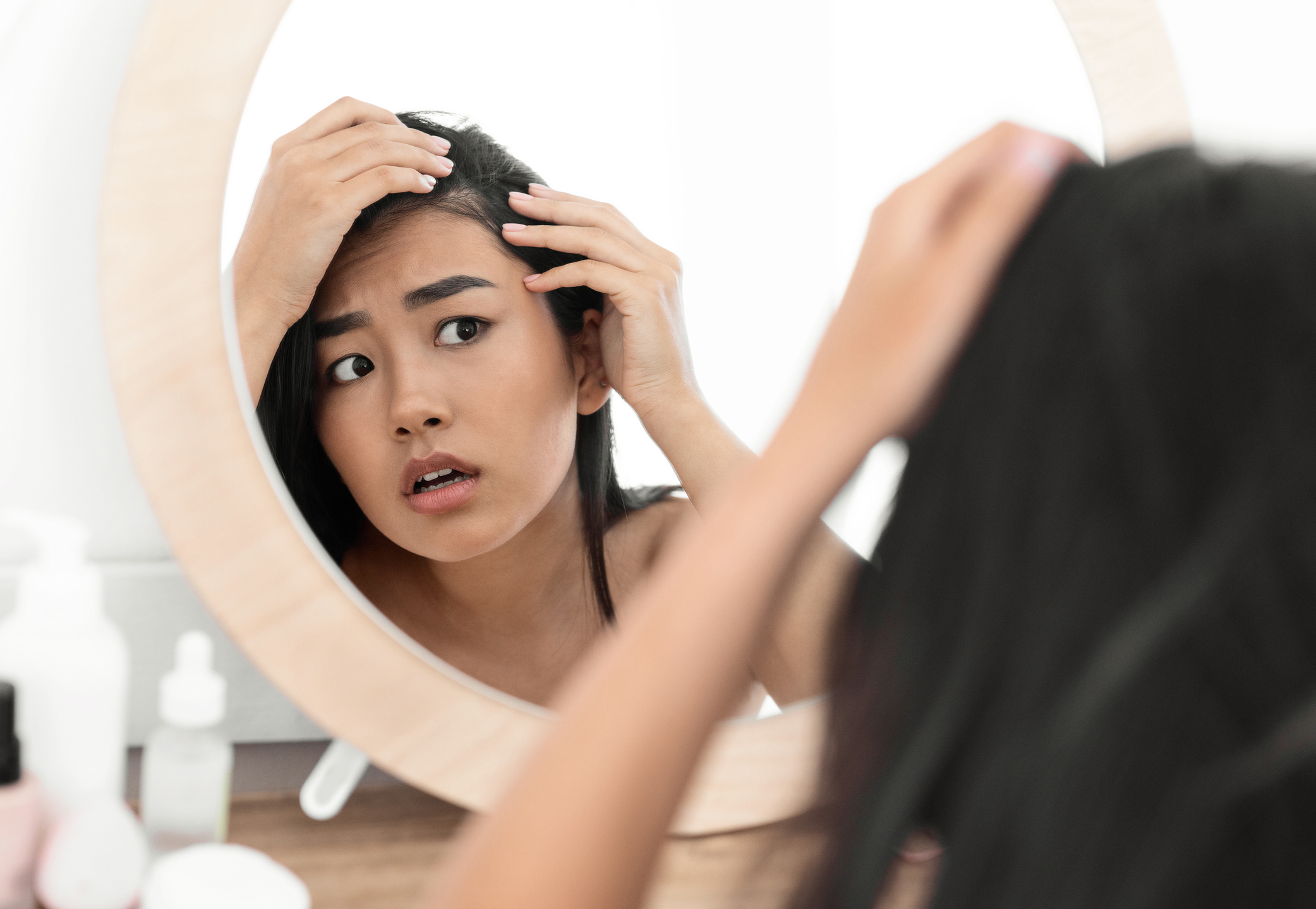 Woman looking closely at her scalp in a mirror, parting her hair with her hands and appearing concerned about hair loss